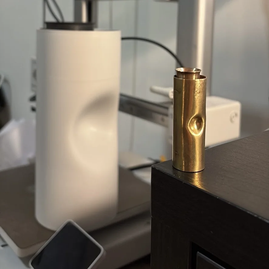 Small brass cylinder prototype with a dimpled detail, next to a 3D printed vase