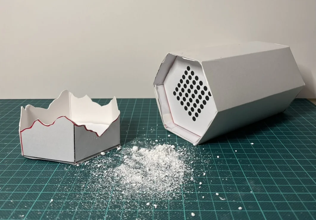 Paper prototype hexagonal carton for climbing chalk, with chalk shaken out through a perforated grid, next to the tray insert